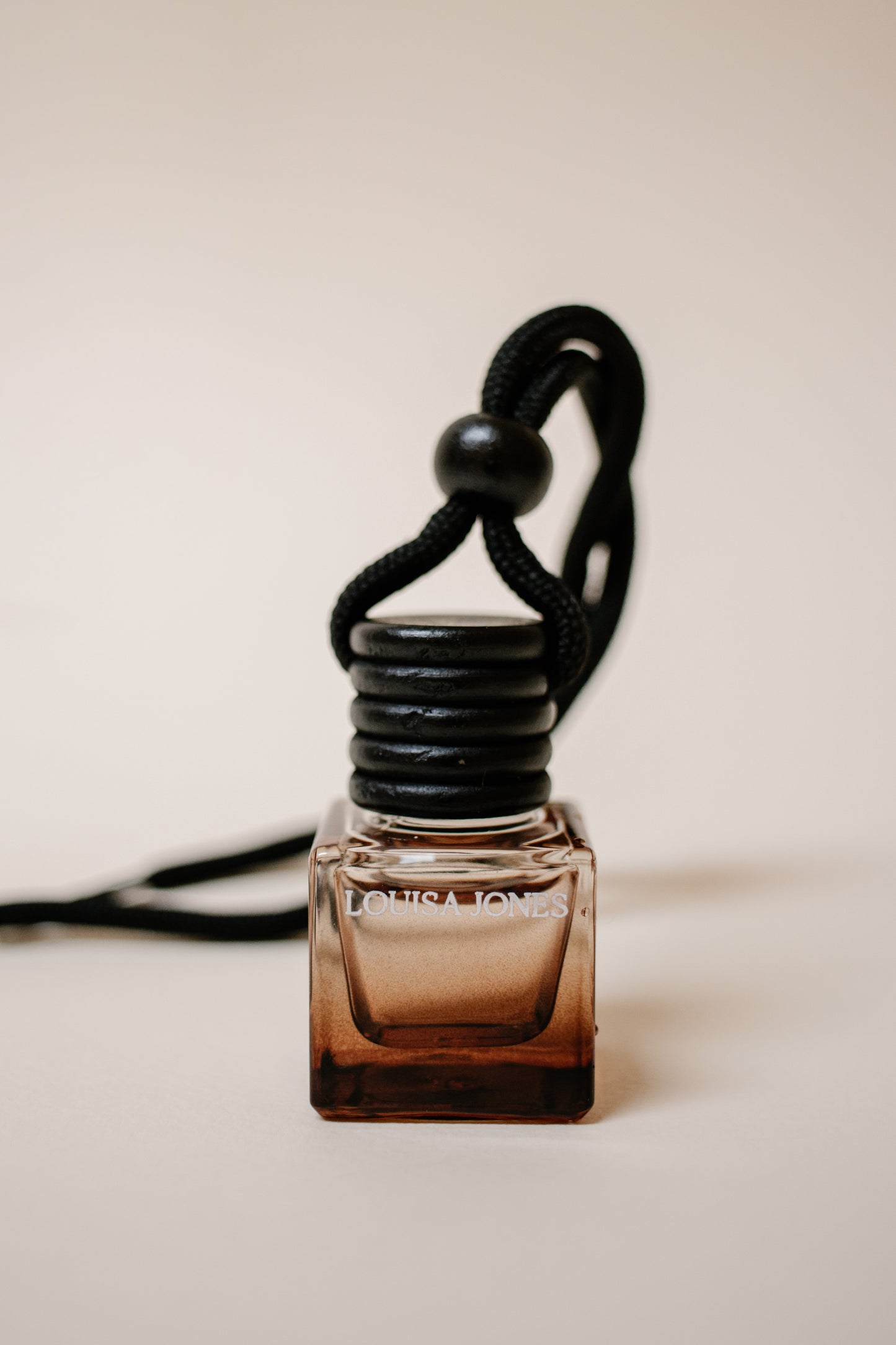 A small square glass car diffuser bottle with a black ribbed cap and a black cord loop, shown on a plain warm beige background. The bottle is amber-brown at the bottom and clear near the top, with the brand name 'LOUISA JONES' printed on the front.
