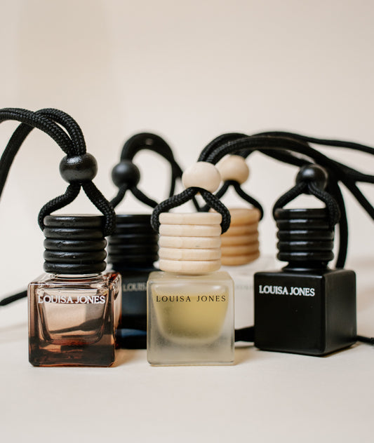Louisa Jones car diffuser bottles with black cords and wooden beads, arranged on a light beige surface against a cream background.