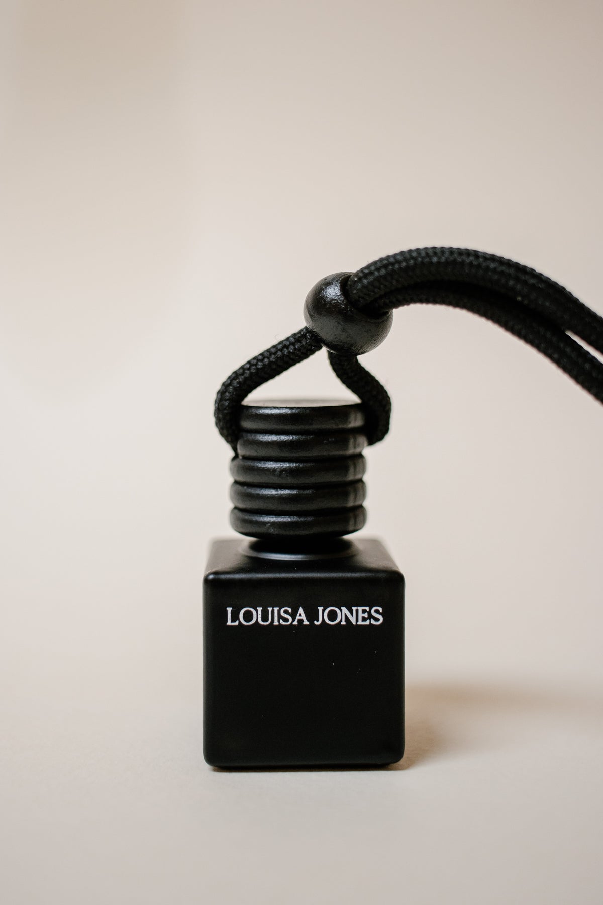 A black square diffuser bottle with a black cord loop and a ribbed black cap, featuring the white text 'LOUISA JONES' on the front, set against a plain beige background.