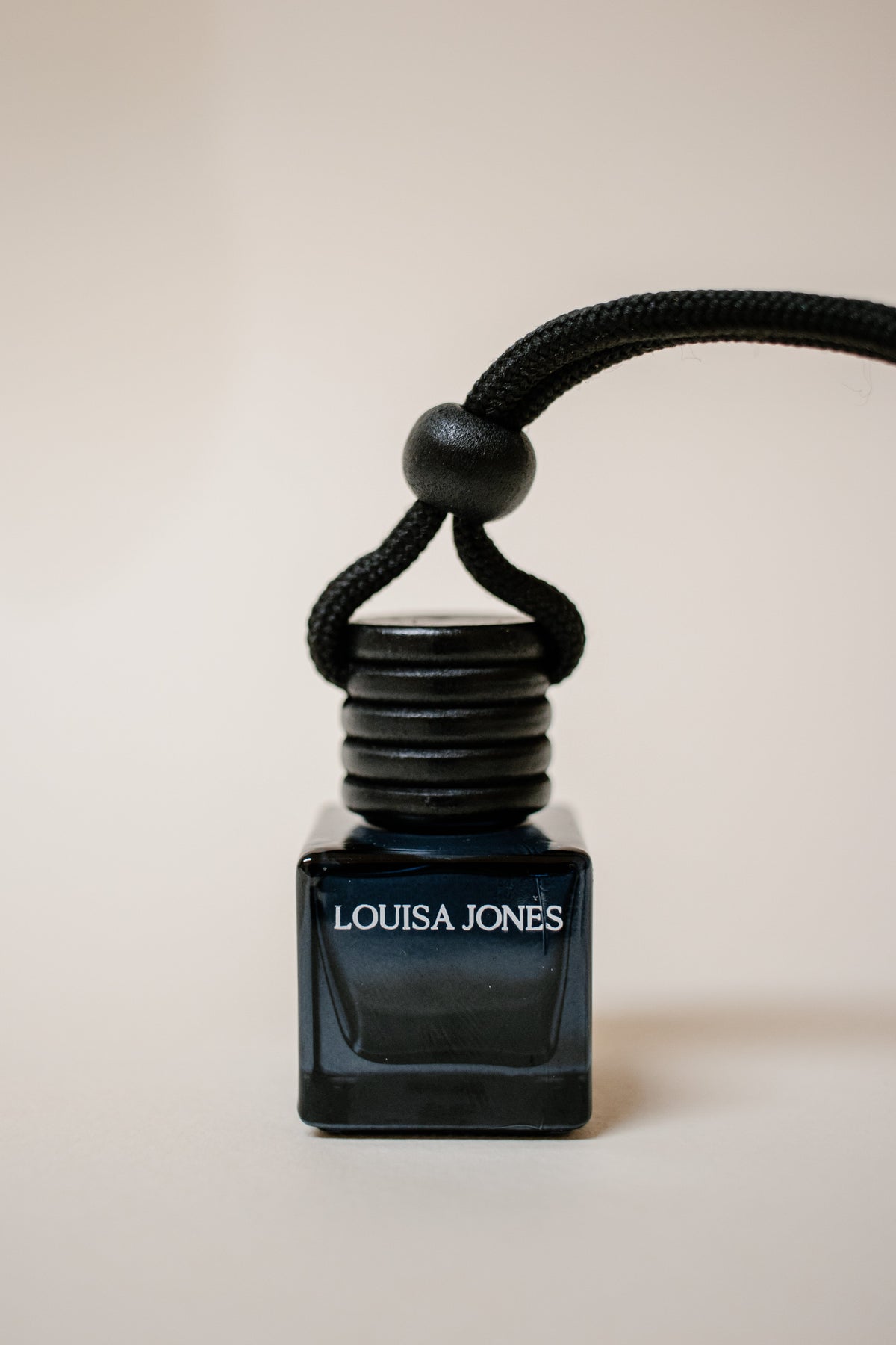 A black glass car diffuser bottle with a black cord, featuring the white LOUISA JONES brand name on the front, set against a solid beige background.
