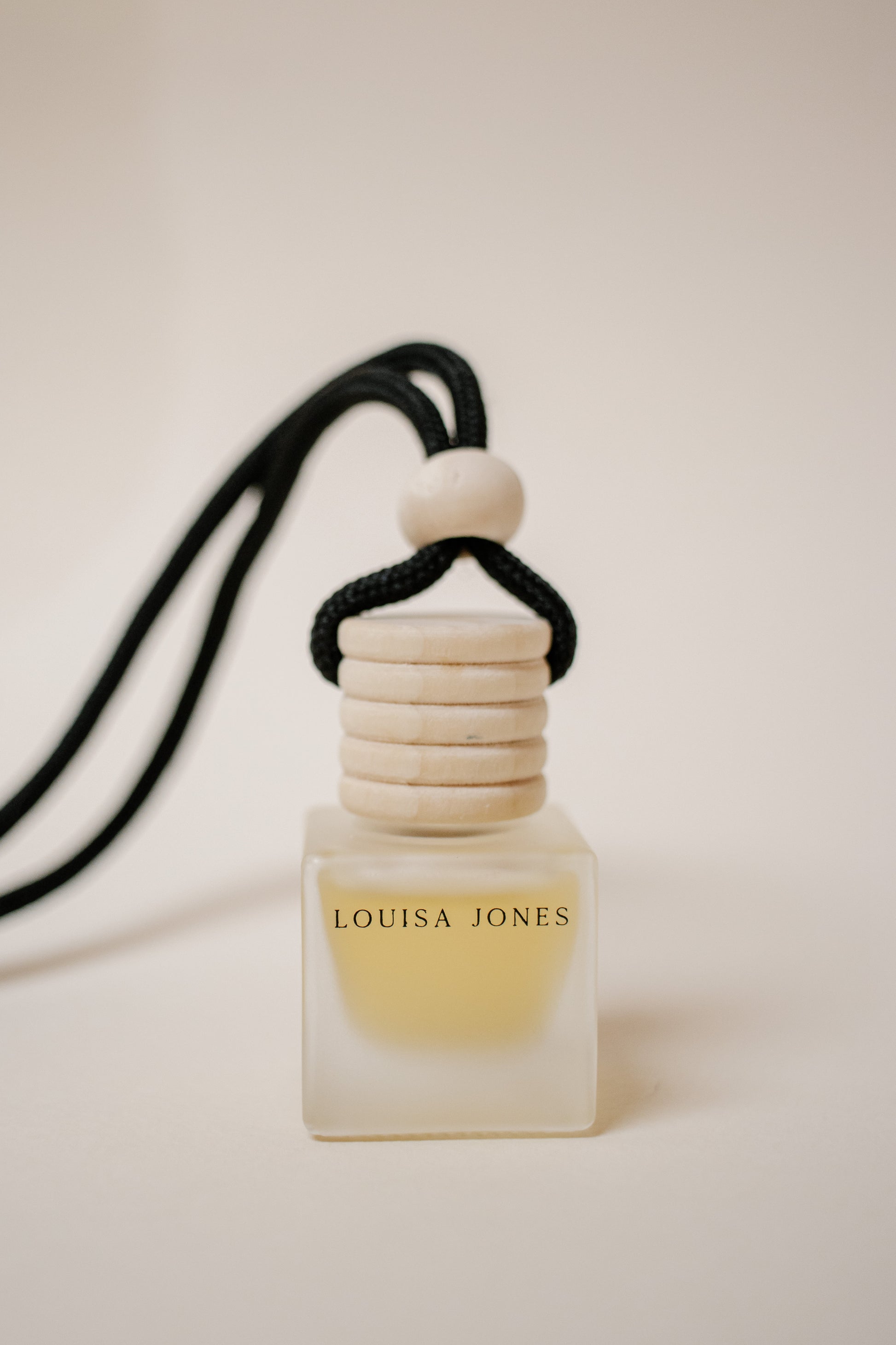 A small square frosted glass car diffuser bottle with a black cord and light wood cap, labeled LOUISA JONES, on a warm off-white background.