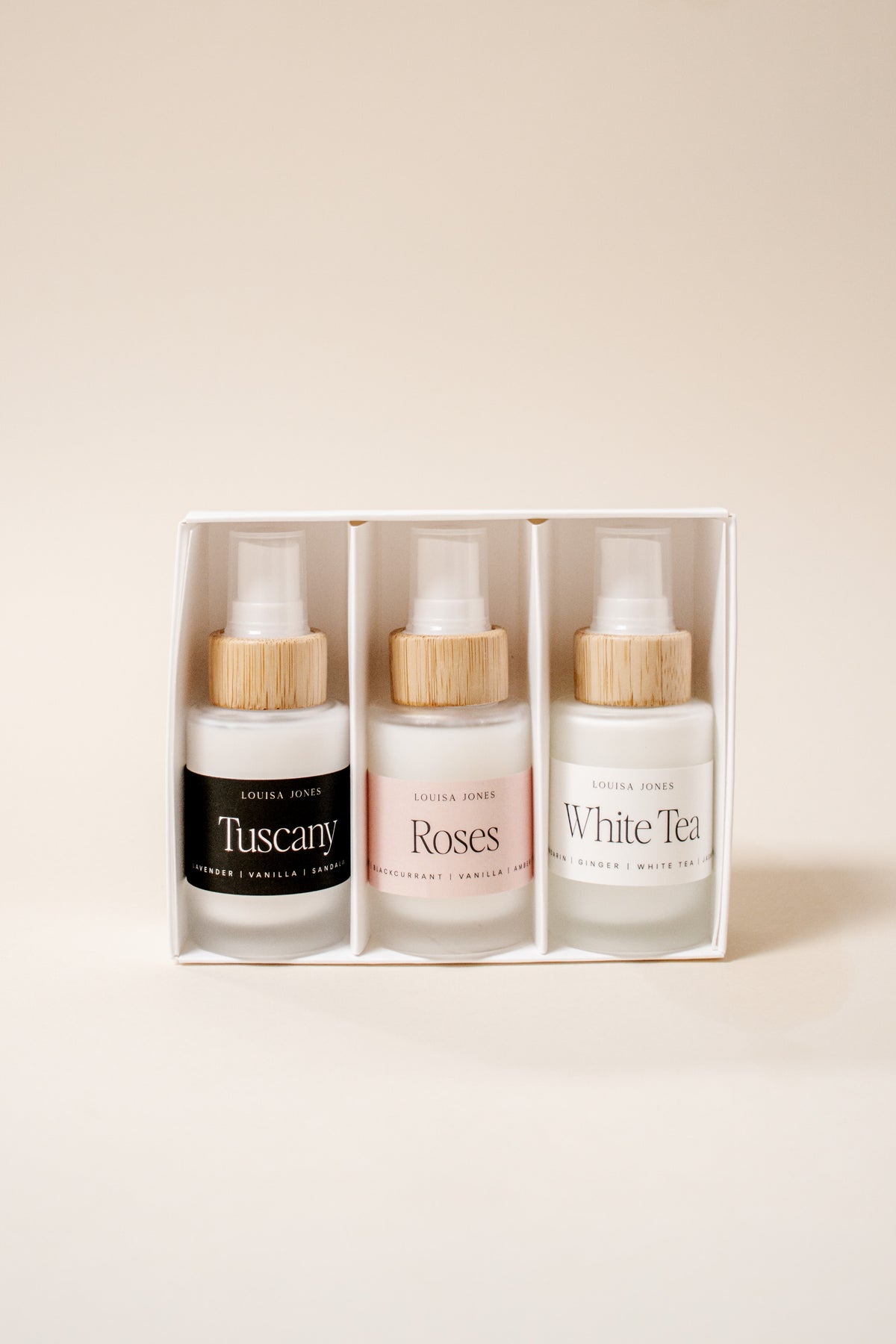 A boxed set of three spray bottles from the brand 'Louisa Jones' with bamboo caps, displayed in a white rectangular box against a warm beige background. The bottles are labeled 'Tuscany', 'Roses', and 'White Tea'.