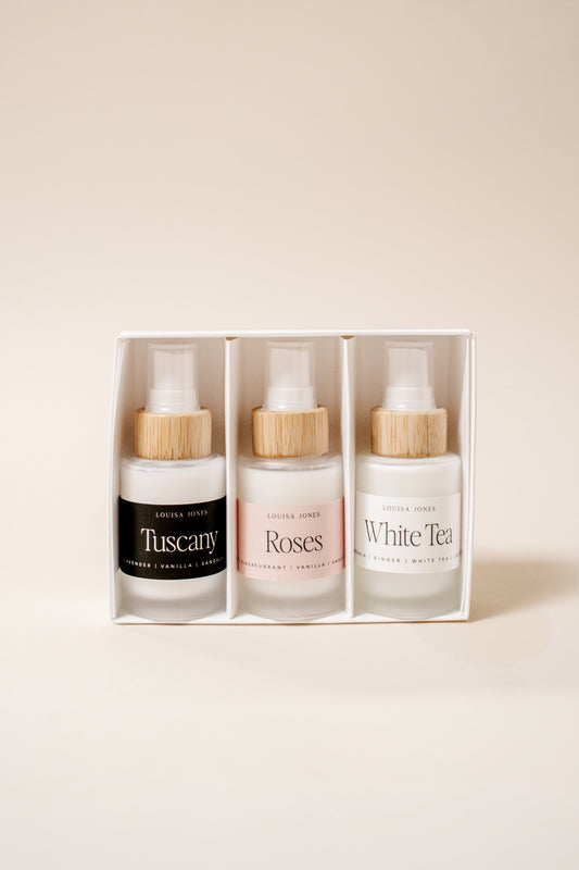 A boxed set of three spray bottles from the brand 'Louisa Jones' with bamboo caps, displayed in a white rectangular box against a warm beige background. The bottles are labeled 'Tuscany', 'Roses', and 'White Tea'.