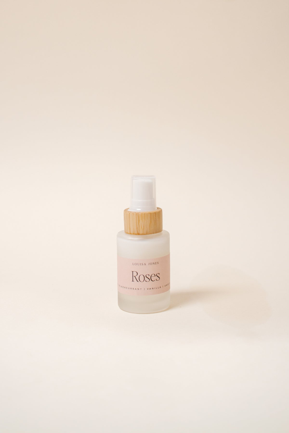 A small frosted glass spray bottle labeled 'Roses' with a light wood-toned collar and white spray top, centered on a warm off-white background.