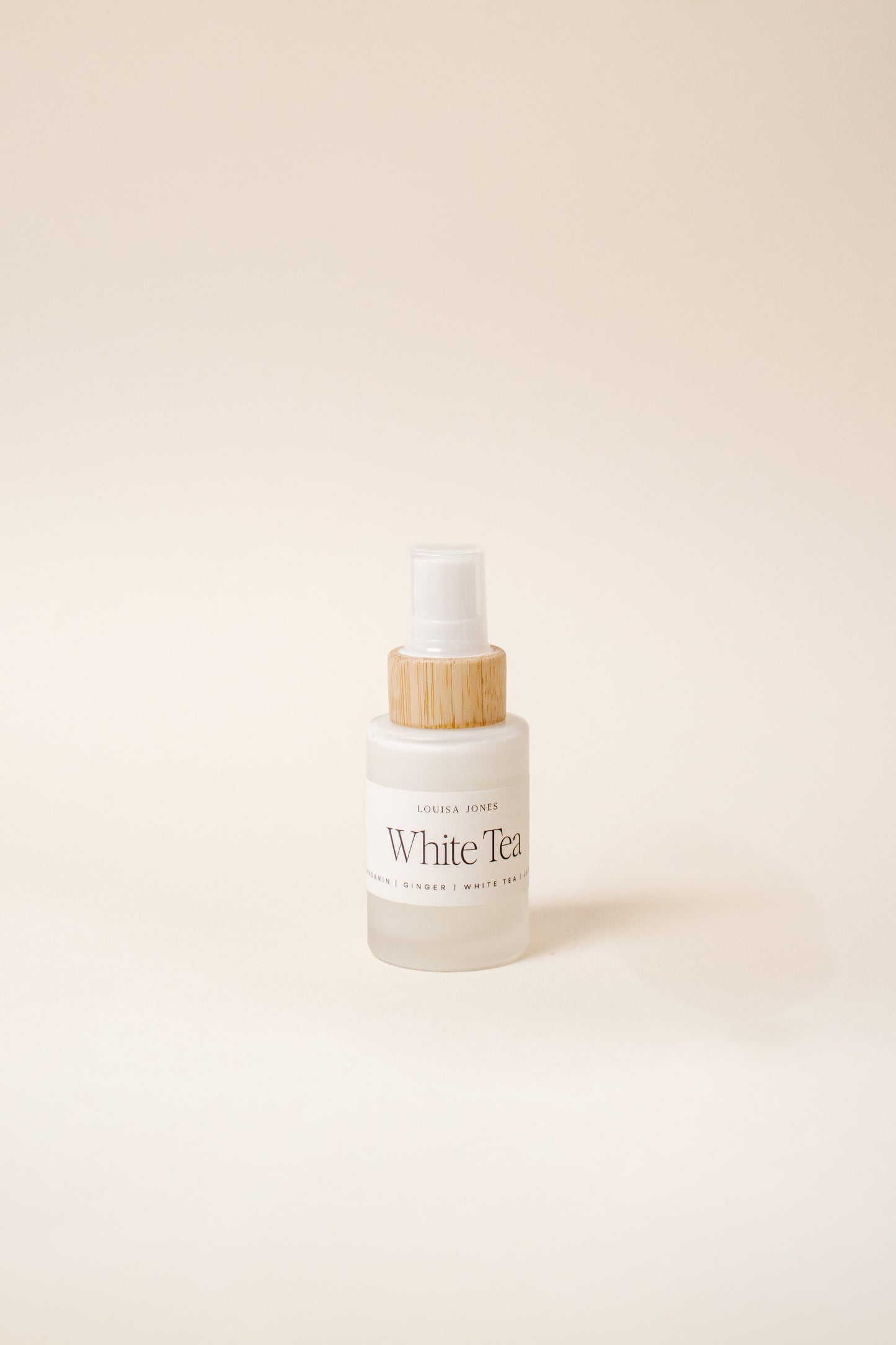 A frosted glass spray bottle labeled 'White Tea' with a light wood-toned collar and white spray top, centered on a warm cream background.