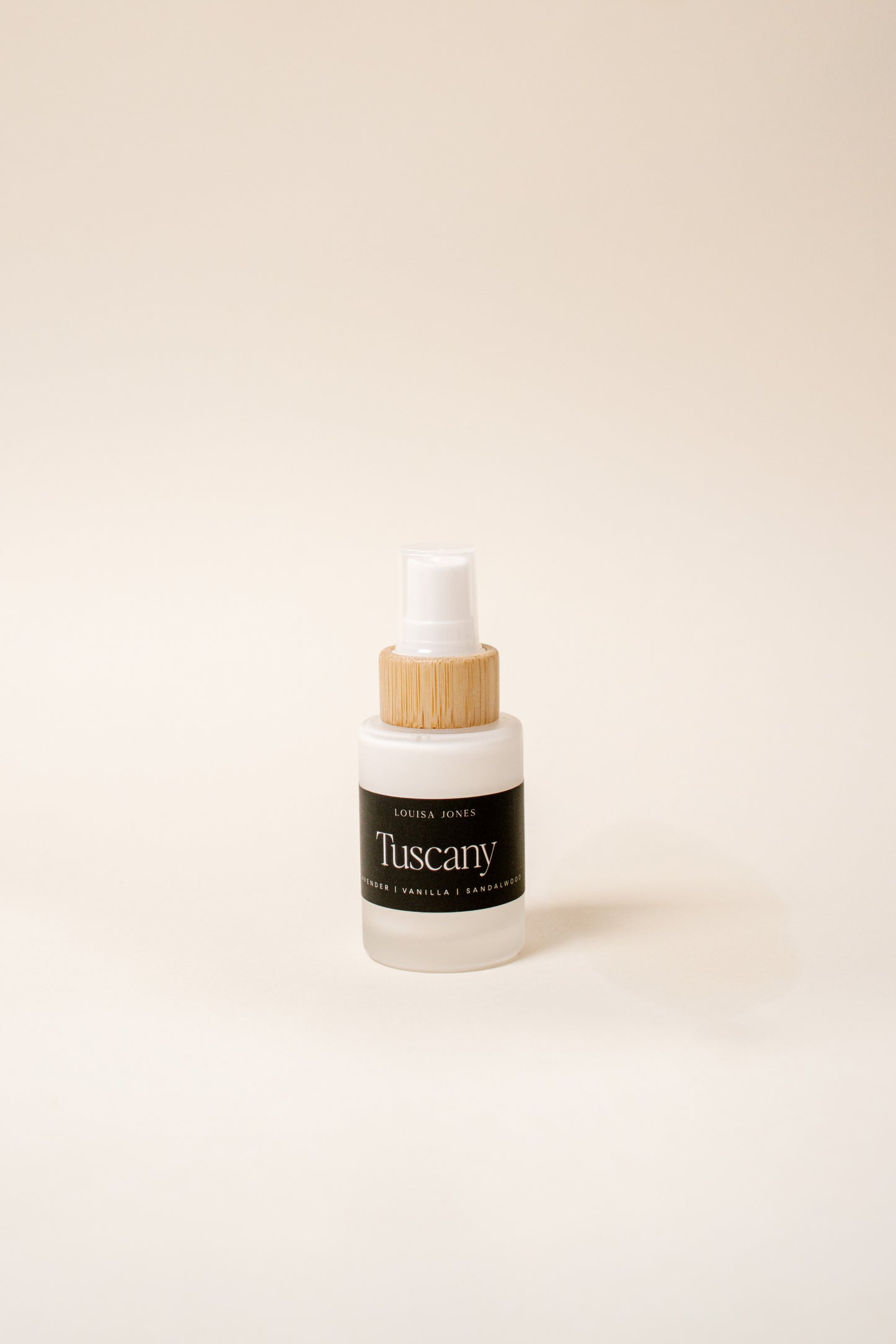 A small frosted glass spray bottle with a white spray top and a bamboo wood collar, featuring a black label that reads 'Tuscany' set against a solid cream background.
