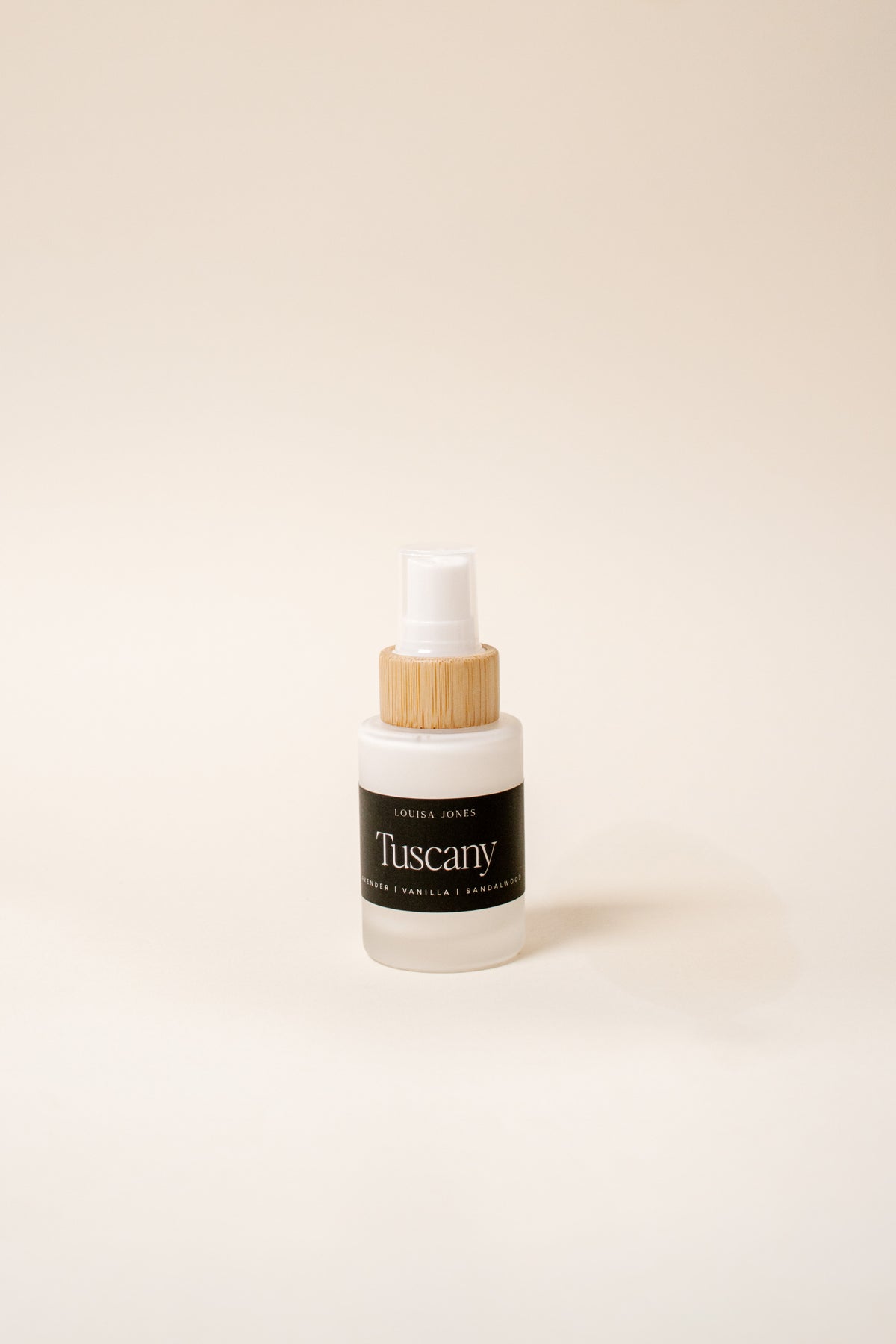 A small frosted glass spray bottle with a white spray top and a bamboo wood collar, featuring a black label that reads 'Tuscany' set against a solid cream background.