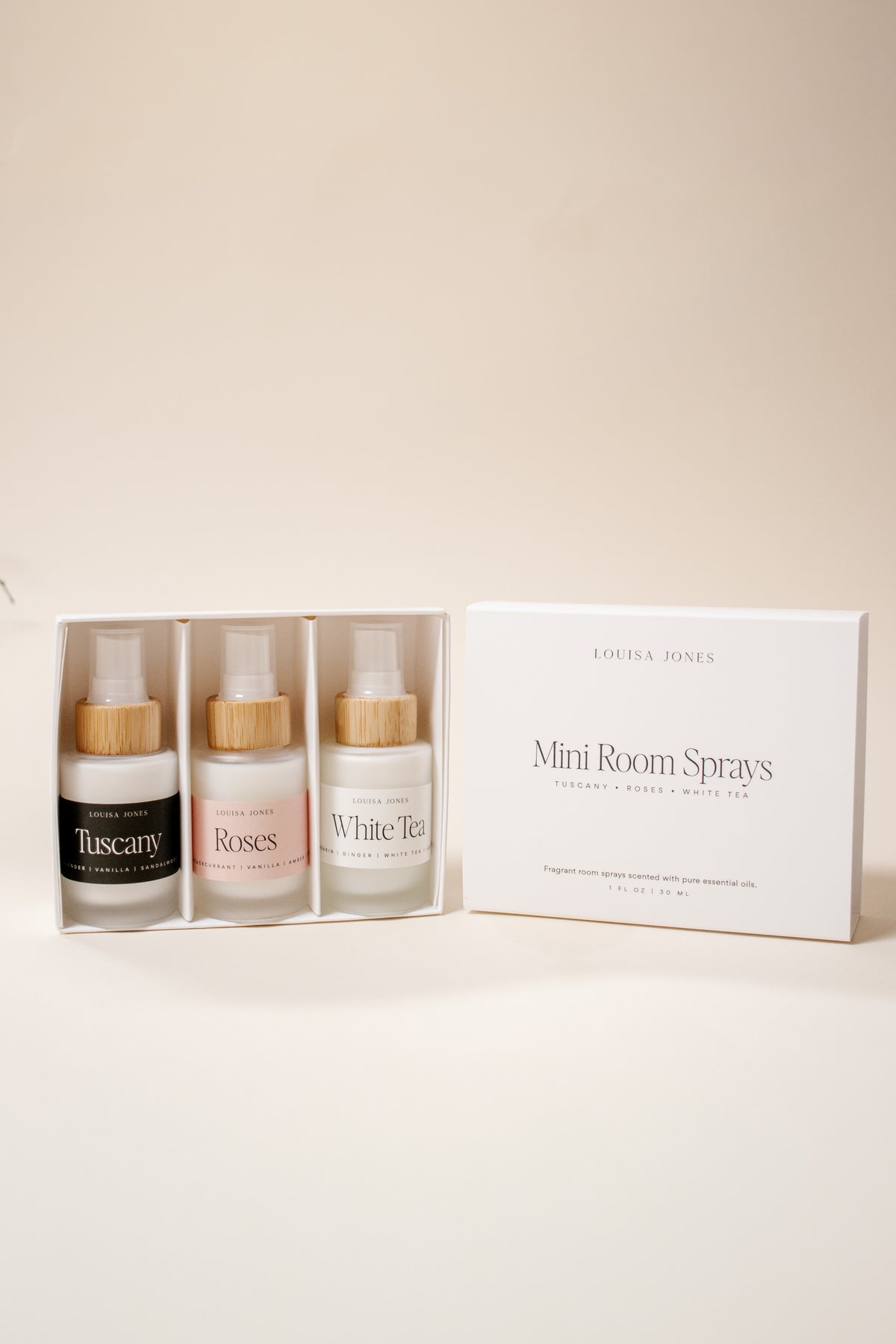 A set of three mini room sprays by Louisa Jones in a white box, shown next to the outer packaging box on a warm beige background. The sprays include Tuscany, Roses, and White Tea, each in a frosted glass bottle with a bamboo cap.